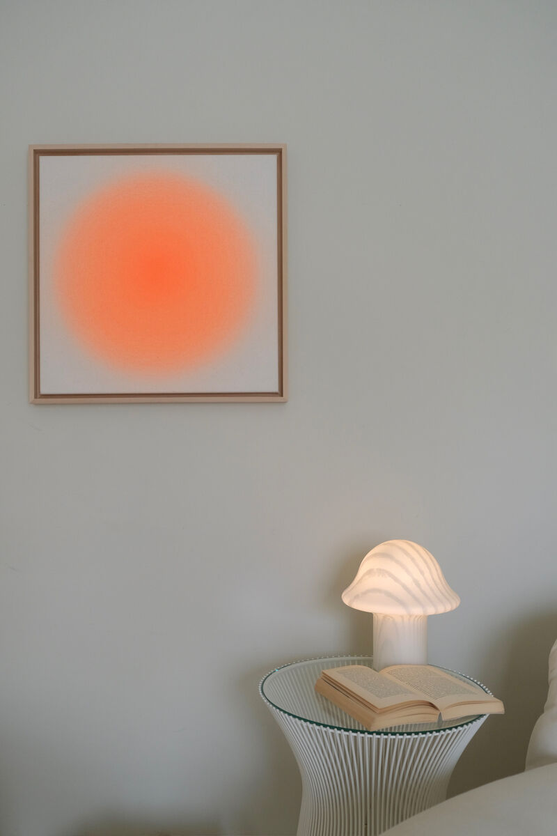 Ein Kunstwerk der ZEIT-Sonderedition Sorbet »Orange I« in 50x50cm Größe hängt an einer weißen Wand. Das Bild hat eine zentrale, orangefarbene Scheibe, die sanft zu den Rändern verblasst. Links daneben steht ein runder Tisch mit weißem Drahtgestell und einer geöffnete Buch mit weißem Einband. Ein geschwungener, weißer Lampenschirm beleuchtet den Tisch.