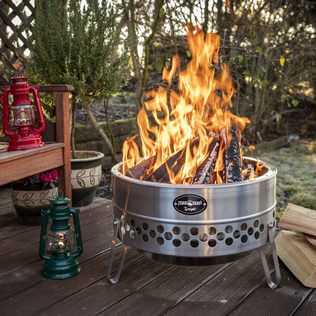 Eine brennende Feuerschale auf einer Holzterrasse, umgeben von Pflanzen und Laternen.
