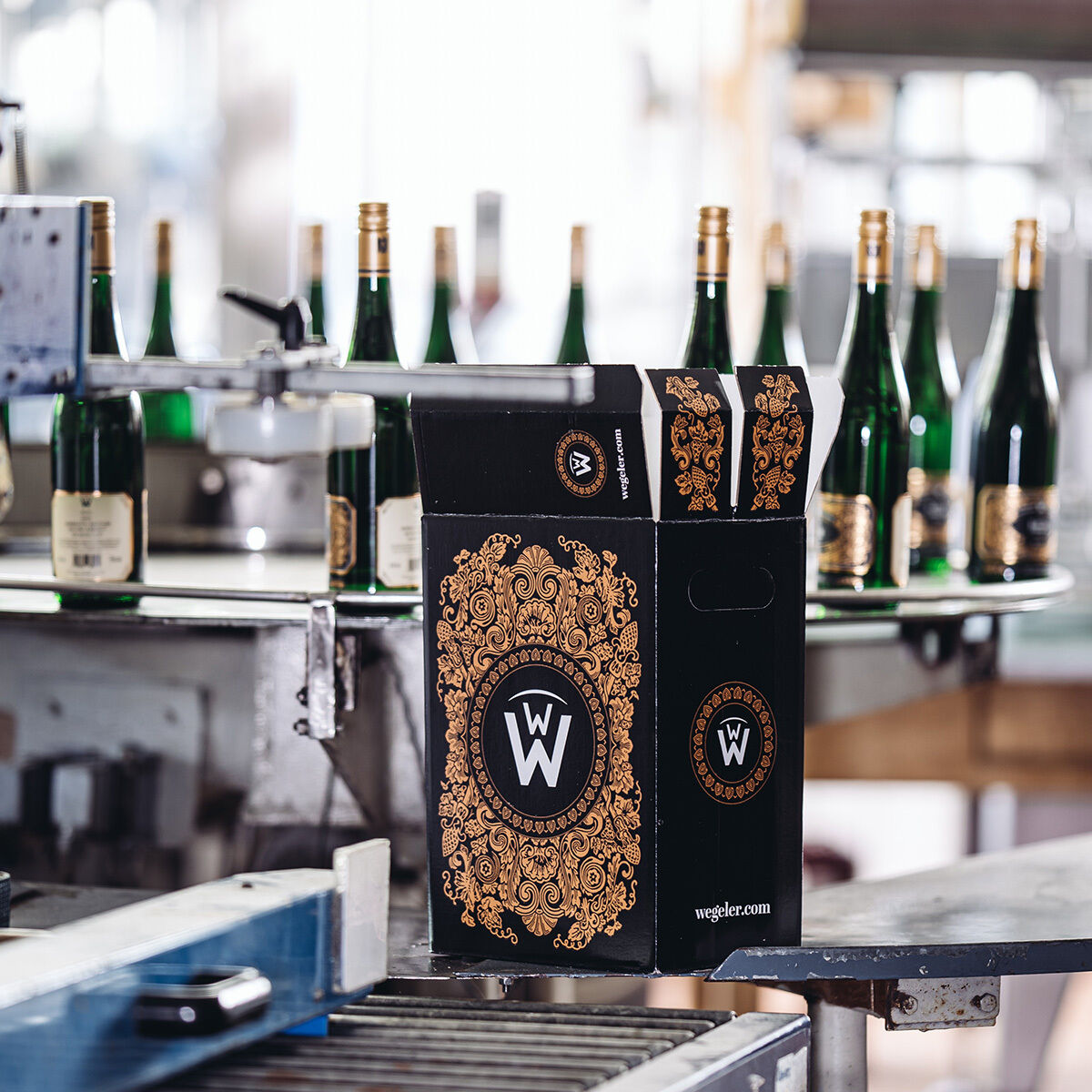 Zu sehen ist ein edles, schwarzes Weinkarton mit goldenen Ornamenten und dem Logo der Weingüter Wegeler, davor eine moderne Flaschenabfüllanlage mit mehreren grünen Weinflaschen. Der Karton trägt das Label der Weinedition »Beethoven«. Die Szene wirkt hochwertig und industriell.