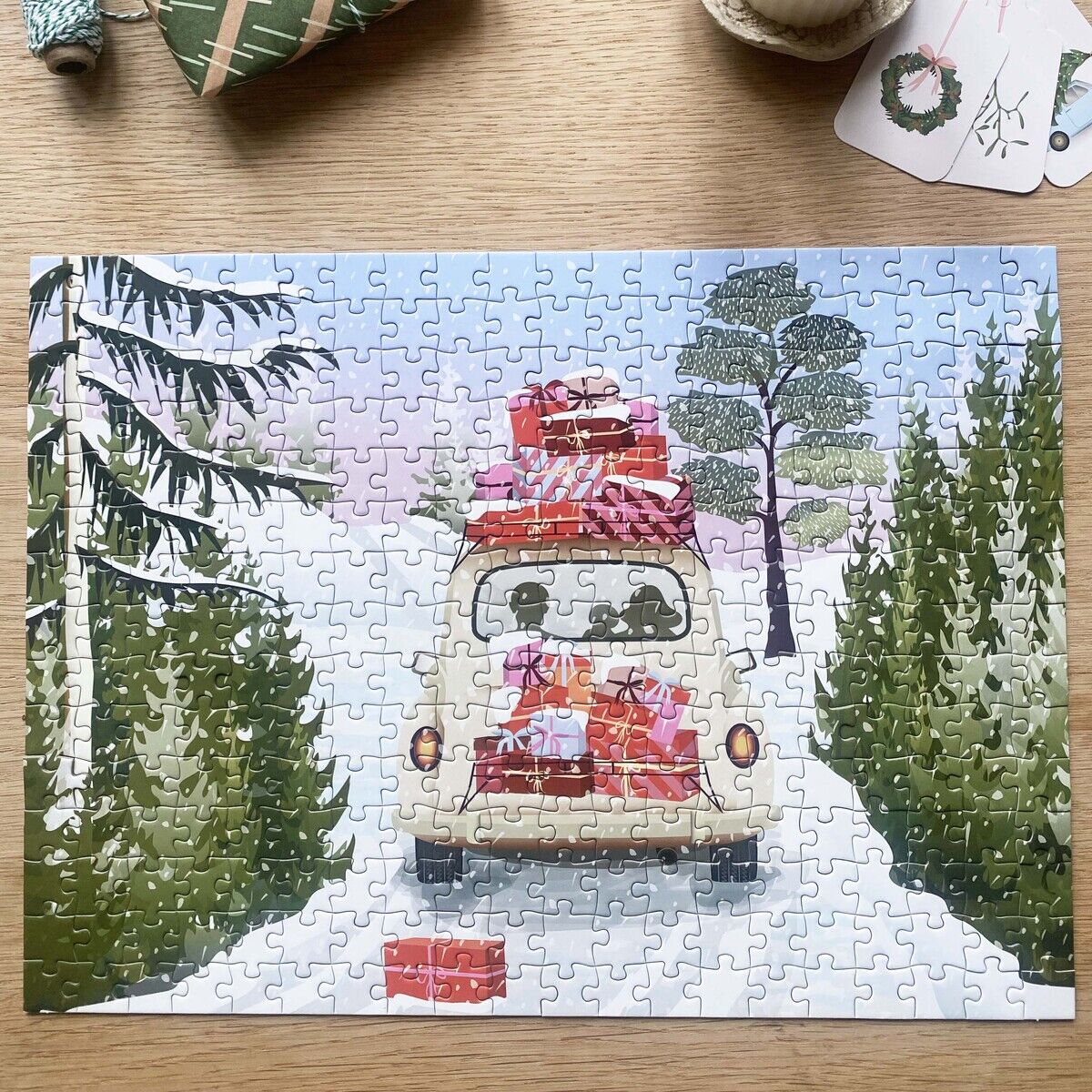 Das 300-teilige Puzzle »Alles gepackt« von VISSEVASSE zeigt ein Auto, das durch eine winterliche Landschaft fährt, auf dessen Dach und Heck viele bunte Geschenke gestapelt sind. Umgeben von verschneiten Tannen, Geschenken, Kerze und Geschenkpapier auf hölzernem Tisch.