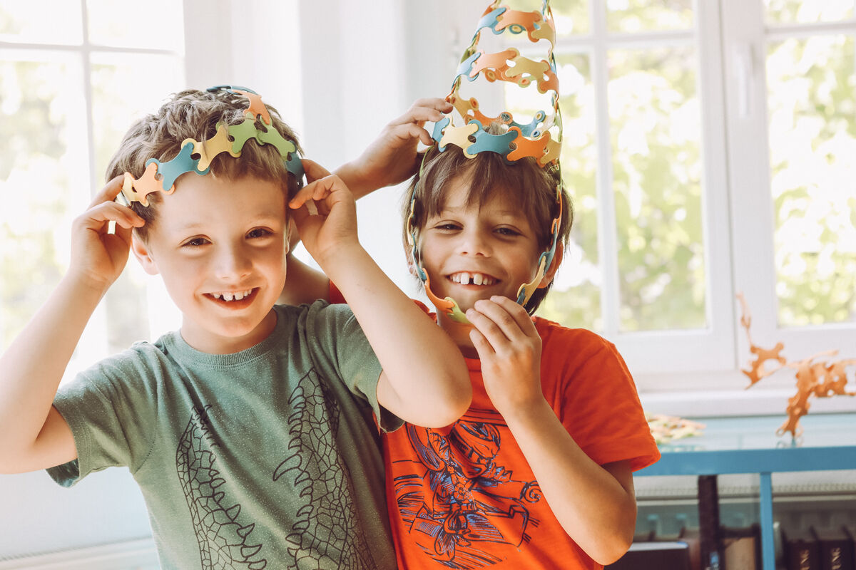 Zwei lachende Jungen, die mit dem Spiel 'Binabo' farbenfrohe Hüte und Schmuckstücke aus Plastikmodulen gebastelt haben. Im Hintergrund ein heller Raum mit großen Fenstern.