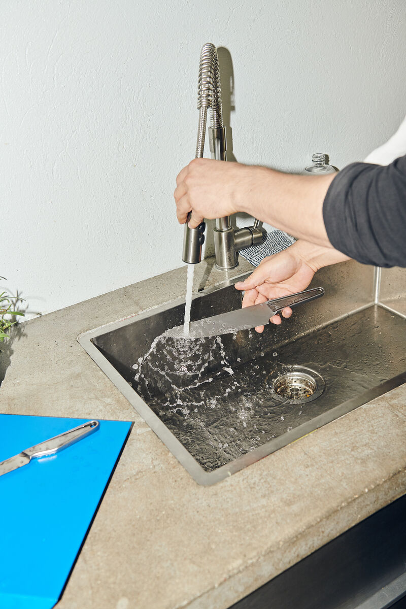 Eine Person spült ein Küchenmesser aus rostfreiem Stahl unter einem Wasserhahn in einer Edelstahlspüle.