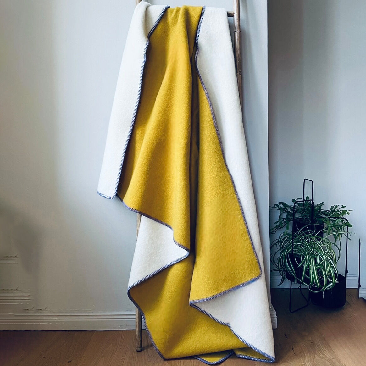 Eine zweifarbige Decke, mit einer gelben und einer weißen Seite, hängt über einer Holzleiter. Daneben steht eine Pflanze auf einem Holzboden in einem modern eingerichteten Raum.