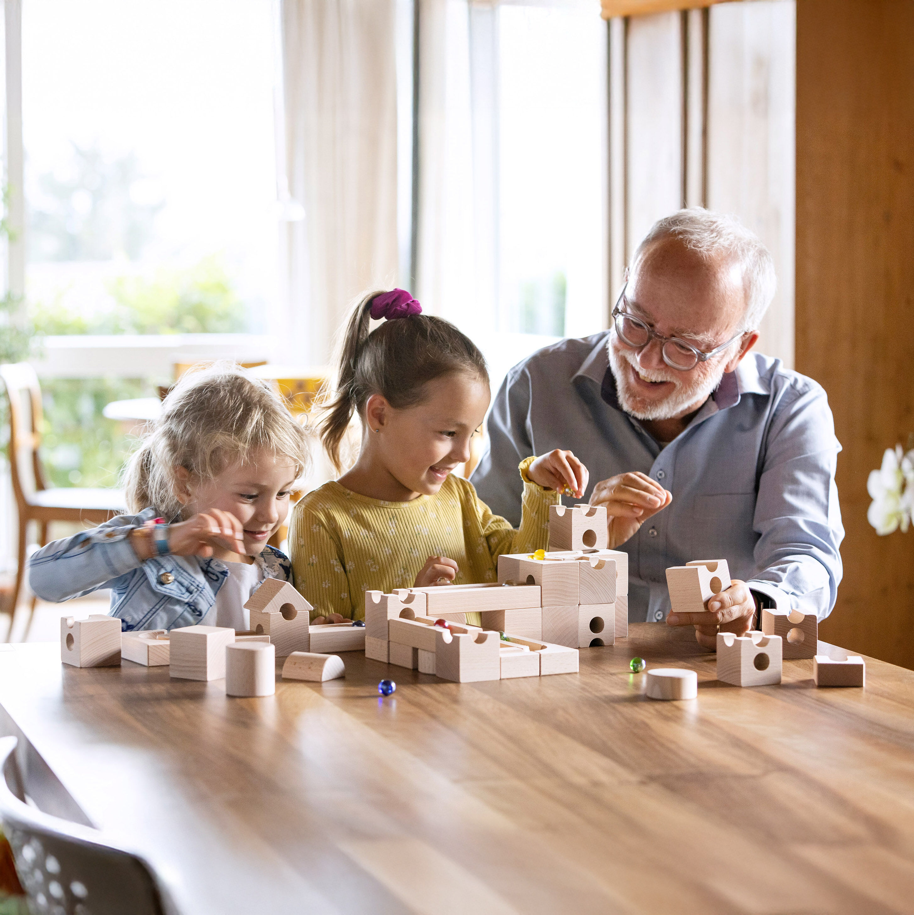 Zwei Mädchen und ein älterer Mann bauen gemeinsam mit dem Cuboro Kugelbahn Set »STANDARD 50 & DAS BUCH« aus hellem Buchenholz eine Murmelbahn auf einem Holztisch. Sonnenlicht fällt durch große Fenster. Die Szene wirkt einladend und kreativ.