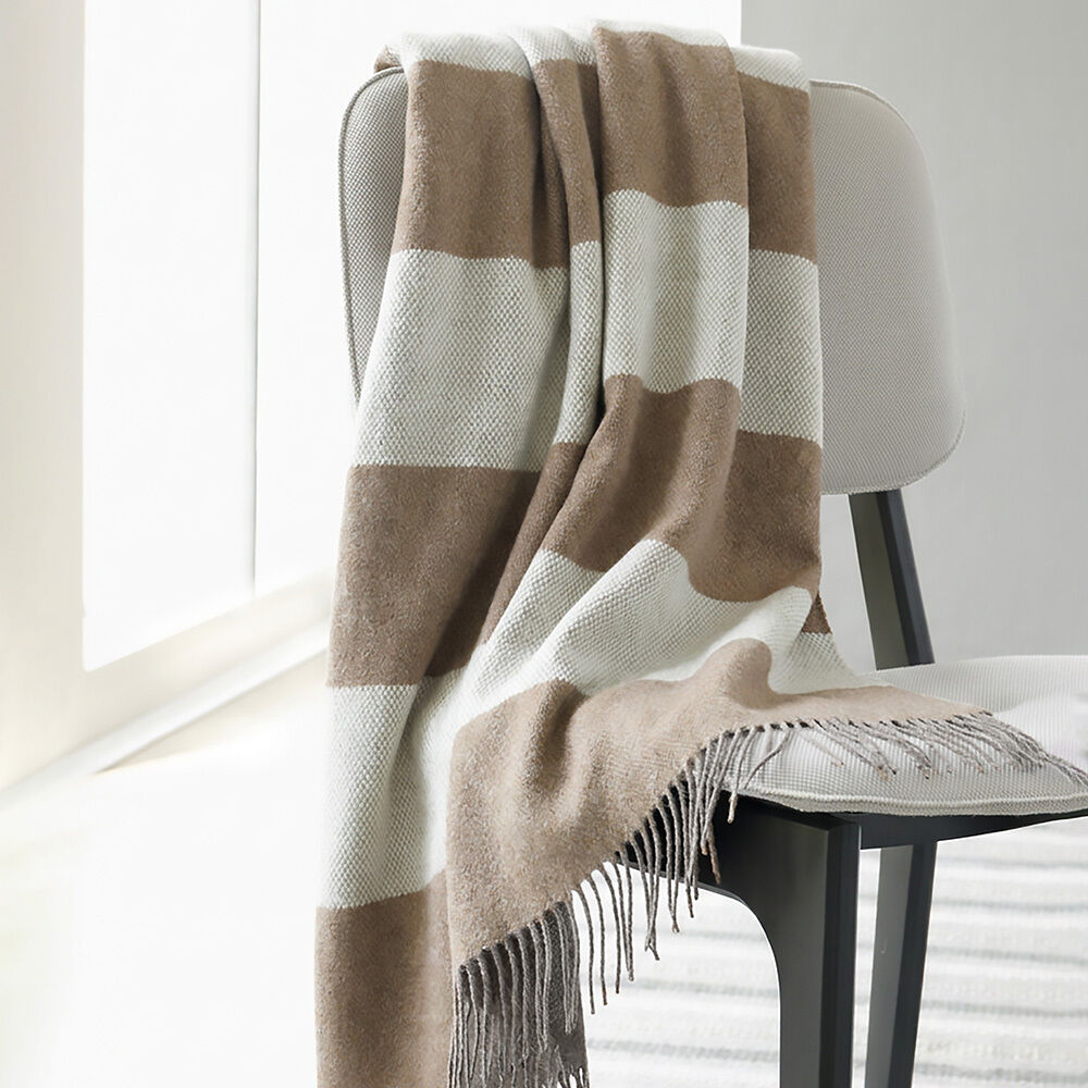 Eine edle Merinodecke »Johann« von Eagle Products in großem Blockstreifenmuster in Beige und Creme liegt locker über einer modernen, hell gepolsterten Stuhllehne. Fransen und die weiche, rustikale Wollstruktur sind gut sichtbar. Helle, einladende Raumatmosphäre.