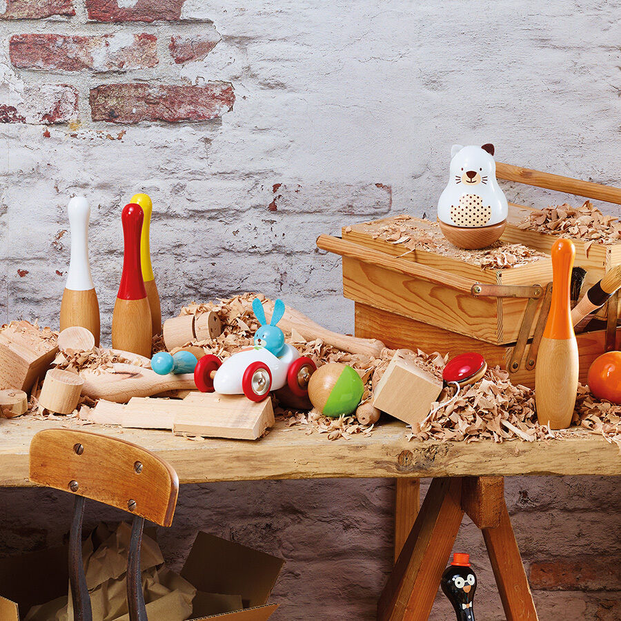 Ein Kegel-Set aus Holz mit verschiedenen bunt bemalten Figuren und Kegelstiften, auf einem Holztisch mit Spänen dekoriert. Im Hintergrund eine gemauerte Wand.