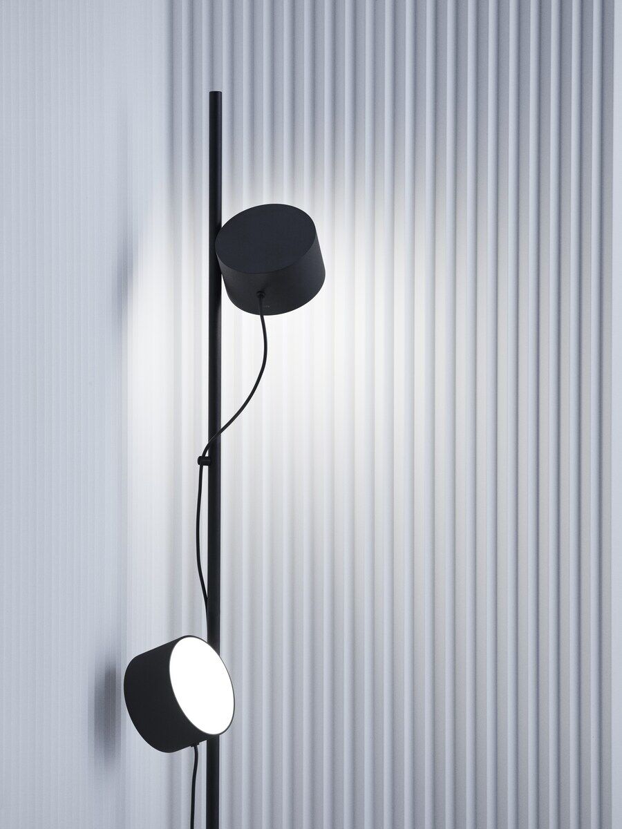 Moderne Bodenlampe »Post« mit minimalistischem Design. Die Lampe ist schwarz und hat zwei zylinderförmige Leuchten. Sie steht vor einer wellenförmigen, grauen Wand.