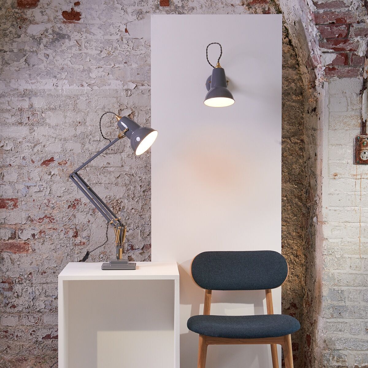 Ein modernes Wohnzimmer mit einer Tischleuchte »Anglepoise Original 1227 Brass Desk Lamp« auf einem weißen Tisch. Die Lampe hat eine graue Farbe mit goldenen Akzenten, spiegelverkehrt von einem passenden Wandlicht. Die Wände zeigen eine Mischung aus freiliegenden Ziegelsteinen und weißer Farbe. Ein grauer Stuhl mit Holzbeinen steht neben dem Tisch.