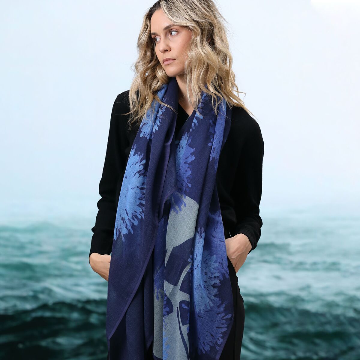 Frau mit langem, blauem Wollschal mit Blättermuster steht vor einer Kulisse mit Meer und Himmel. Der Schal wirkt weich und wärmend und ist als modisches Accessoire für Herbst und Winter dargestellt.