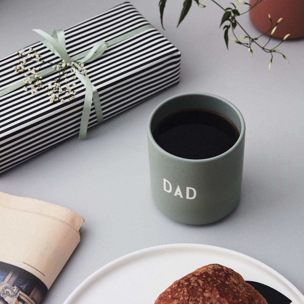 Farblich abgestimmtes Arrangement mit einem grünen Becher mit der Aufschrift ‚Dad‘, einem eingepackten Geschenk, einem Croissant auf einem weißen Teller, einer Zeitung und einem Pflanzendekor auf einem grauen Hintergrund.