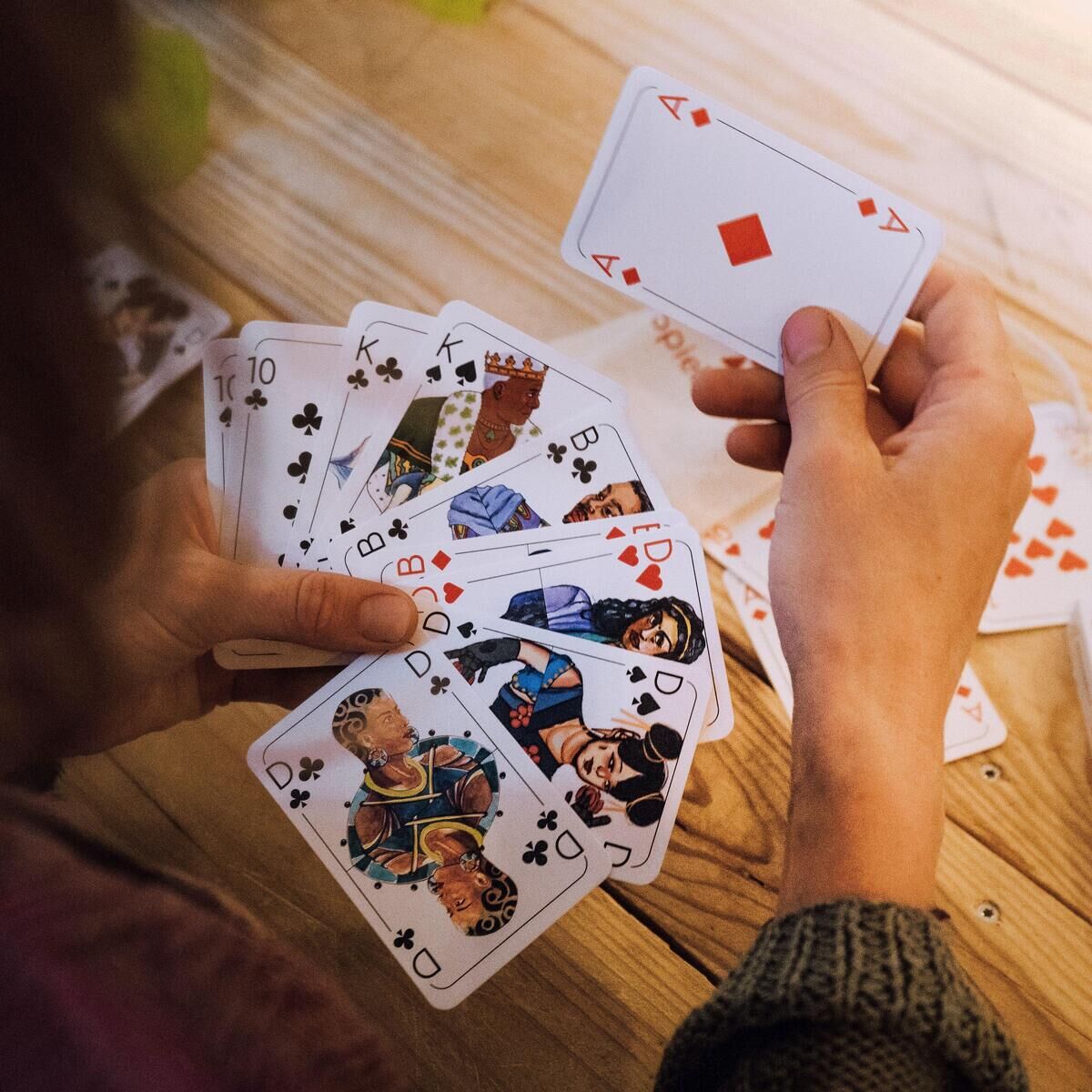 Eine Person hält eine Handvoll Spielkarten für das Kartenspiel 'Doppelkopf'. Die Karten zeigen verschiedene Bilder und Symbole. Der Spieltisch ist aus Holz und weitere Karten liegen darauf verstreut.