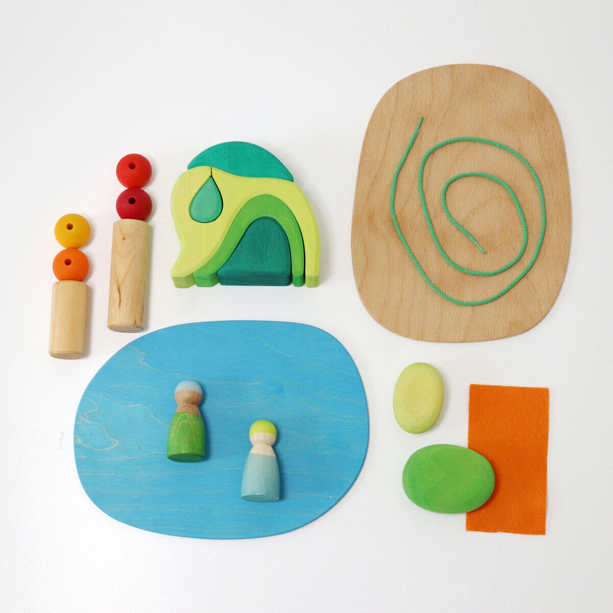 Eine Spielwelt namens »Waldlichtung«, bestehend aus verschiedenen Holzspielzeugen. Zu sehen sind bunte Holzelemente in Grün, Blau, Holzfarben und Orange, darunter Spielfiguren, Platten und Naturformen in Form von Steinen und Pflanzen. Auf hellem Untergrund arrangiert.