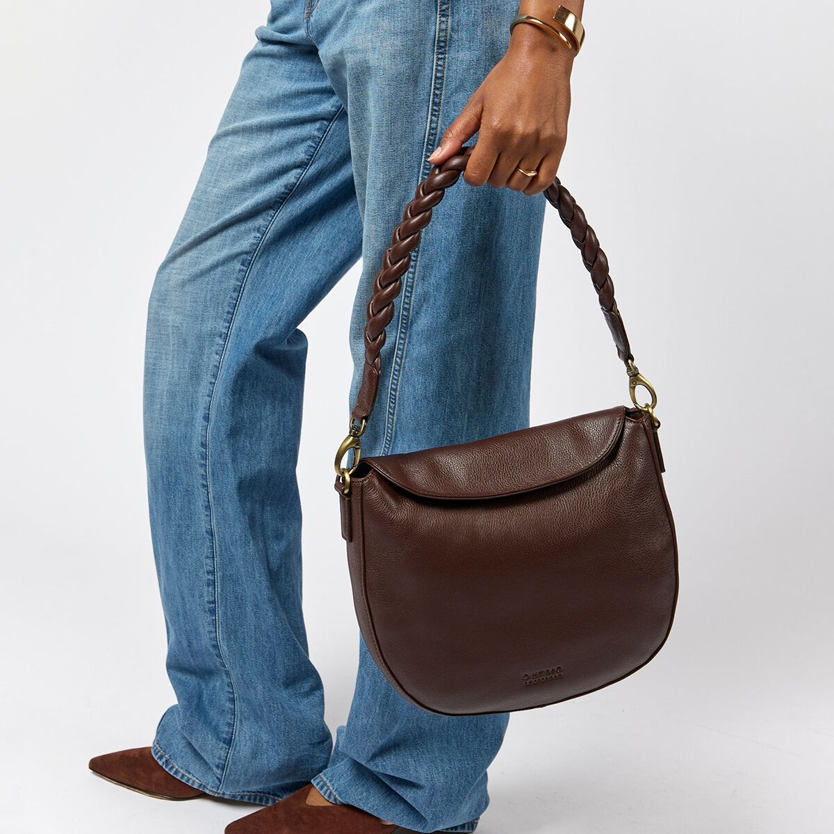 Frau hält eine braune Umhängetasche namens »Lana« aus Lederoptik mit geflochtenem Henkel. Das Modell trägt eine hellblaue Jeans und ein goldfarbenes Armband. Die Tasche wirkt modern und alltagstauglich.