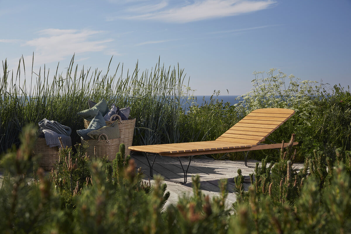 Eine moderne Holzliege von MOLO steht auf einer Terrasse im Freien mit Blick auf das Meer. Im Hintergrund sind hohe Gräser und ein klarer blauer Himmel zu sehen. Neben der Liege stehen Körbe mit Textilien.