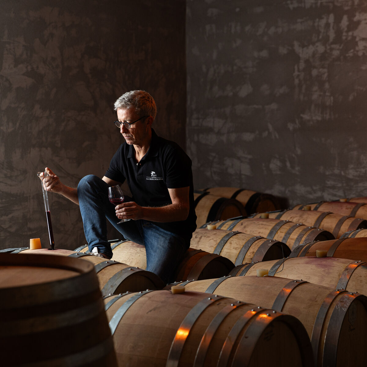 Ein Mann im schwarzen Poloshirt sitzt auf einem Holzfass in einem rustikalen Weinkeller und zieht mit einer Pipette Rotwein aus einem Fass. Um ihn herum stehen mehrere Holzfässer in schummrigem Licht. Das Bild vermittelt einen handwerklichen und traditionellen Eindruck der Weinherstellung.