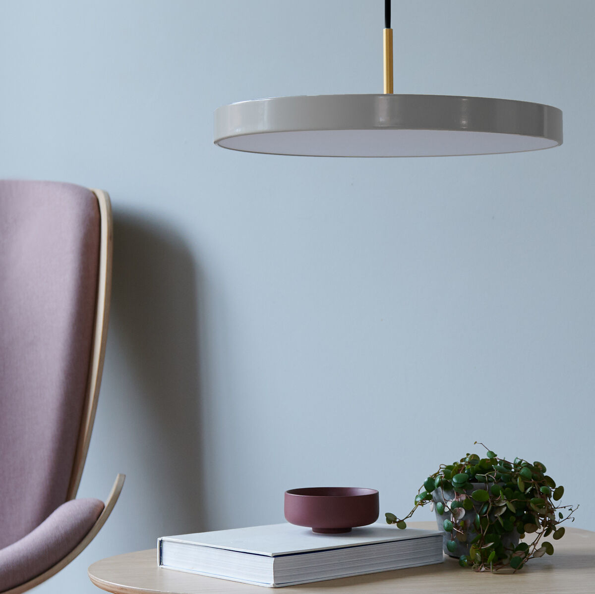 Pendelleuchte »Asteria Medium« mit grauem Lampenschirm und goldenen Akzenten, hängend über einem hellen Holztisch mit einem Buch, eine violette Schale und eine grüne Zimmerpflanze daneben. Im Hintergrund ein beiger Stuhl vor einer hellgrauen Wand.