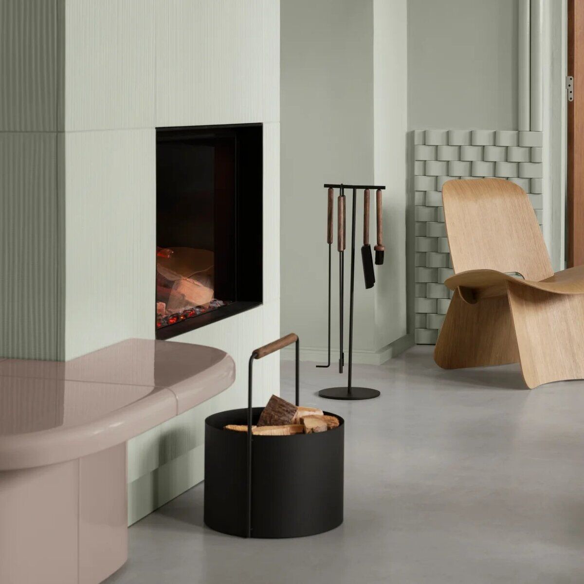 Modernes Kaminbesteck-Set ASHI in Schwarz steht neben einem offenen Kamin in einem minimalistisch eingerichteten Wohnzimmer mit hellen Wänden, einem Holzstuhl und Holzkorb. Die Szene vermittelt eine warme, stilvolle Atmosphäre und eignet sich für Gemütlichkeit in der Winterzeit.