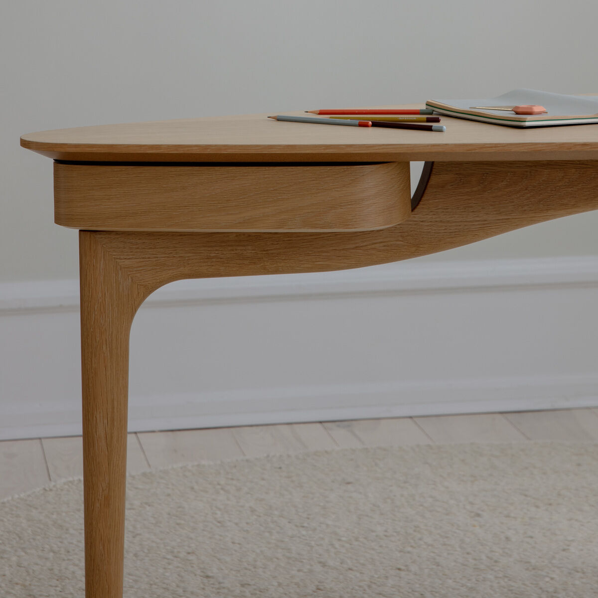Moderner, minimalistischer Schreibtisch aus hellem Holz mit abgerundeten Kanten, darauf liegen einige Stifte sowie Hefte, vor einer hellen Wand und auf einem hellen Teppich stehend.