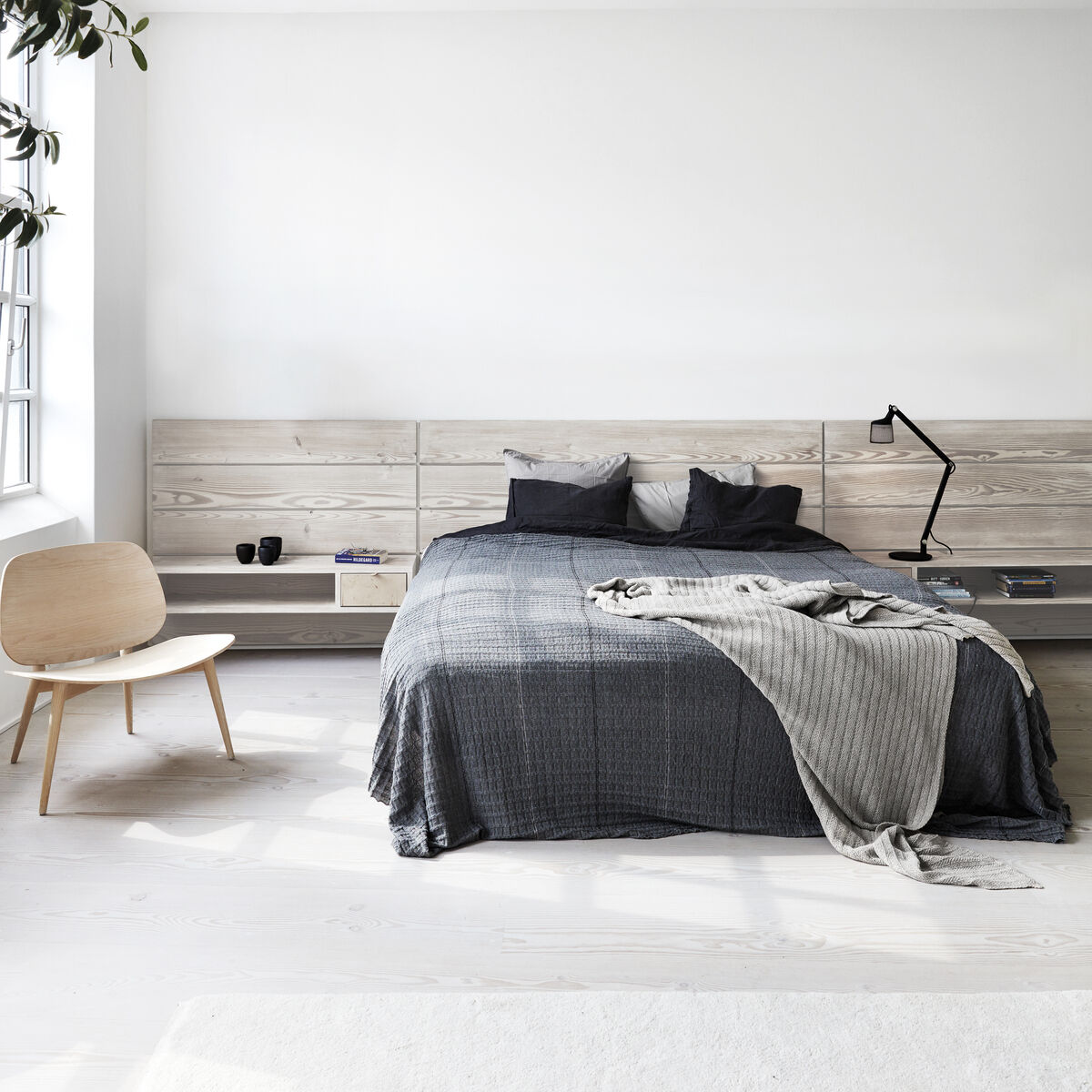 Ein modern eingerichtetes Schlafzimmer mit weißer Wand und hellem Holzboden. Ein graues Bett mit passendem Überwurf, eine minimalistische Schreibtischlampe von Vipp, ein hölzerner Stuhl und ein großes Fenster sind zu sehen.