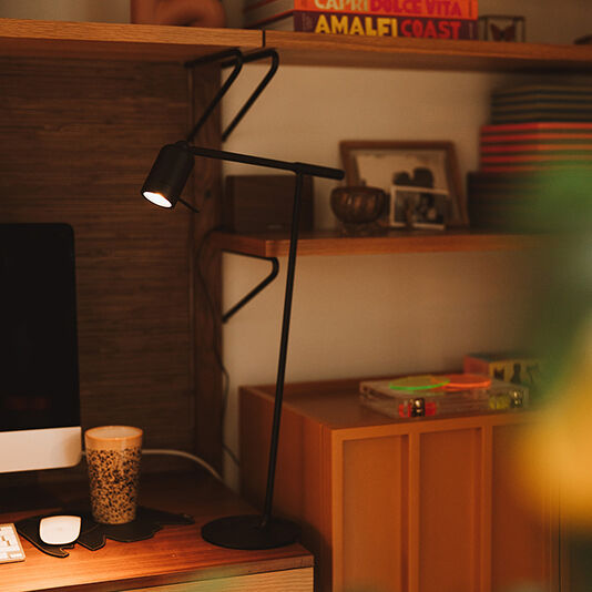 Ein stilvolles Home-Office-Setup mit einem modernen Schreibtisch, einem Monitor, einer Schreibtisch-Lampe Leuchte »ONE« und Bücherregalen im Hintergrund. Die Szene ist warm beleuchtet und strahlt Gemütlichkeit aus.