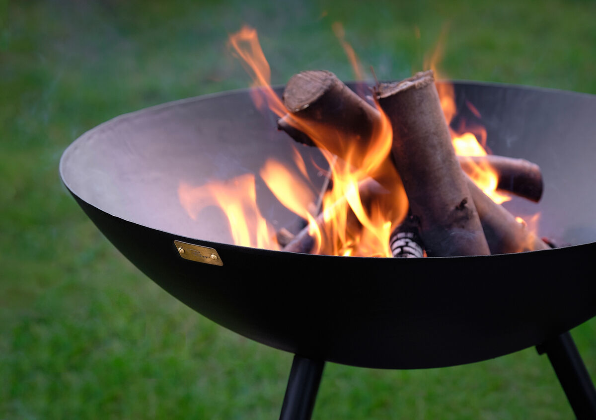 Eine schwarze Feuerschale der Marke »FLAMES« mit brennendem Holz, fotografiert im Freien auf grünem Rasen.