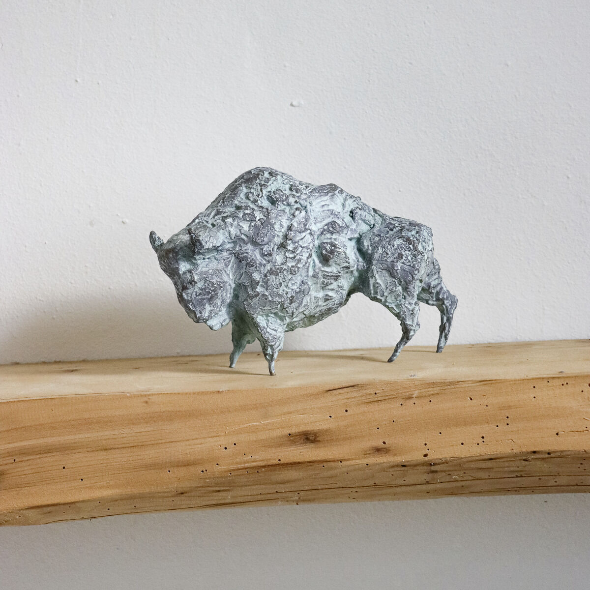 Bronzeskulptur eines Bisons von Michael Jastram auf einem naturbelassenen Holzbrett vor weißer Wand. Die Skulptur zeigt detailreiche Strukturen und einen grünlichen Patina-Effekt.