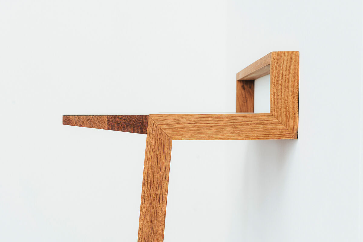 Ein modernes Stehpult aus Holz mit minimalistischer Gestaltung. Es besteht aus einer geometrisch geformten Holzstruktur, die an einer weißen Wand befestigt ist.
