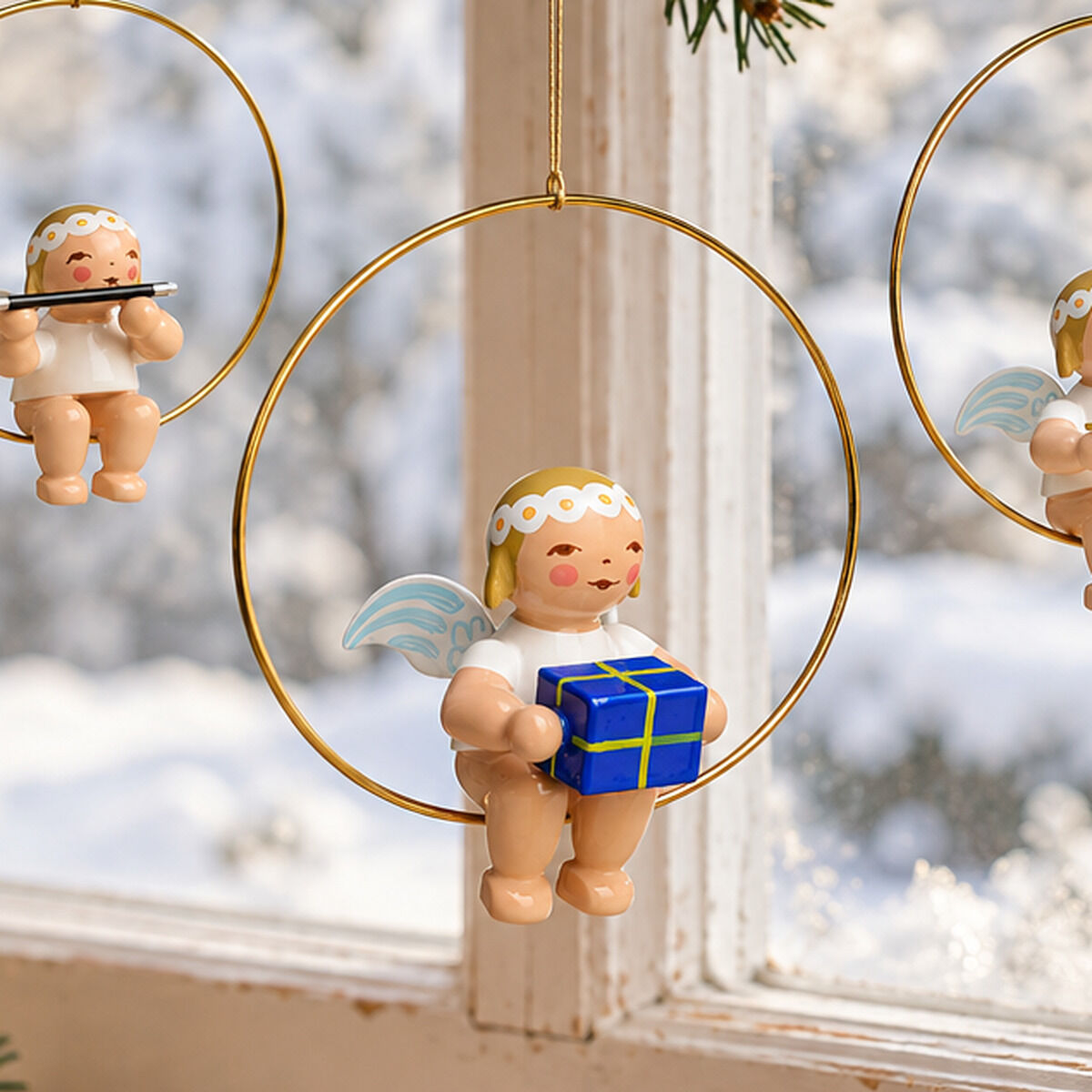 Fünf handgefertigte Holzengel von Wendt & Kühn hängen in goldenen Ringen an grünen Tannenzweigen vor einem verschneiten Fenster. Jeder Engel hält ein anderes Accessoire, zum Beispiel ein Geschenk oder Musikinstrument. Nebenan brennt eine Kerze.