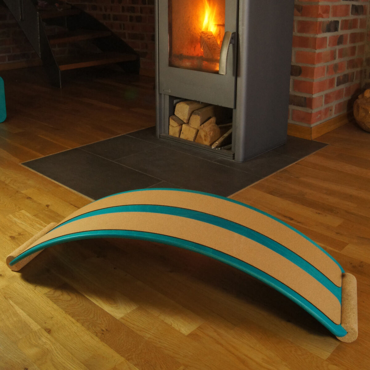 Ein Balance Board aus Holz mit türkisfarbenen Streifen liegt auf einem Holzboden neben einem Kaminofen, der ein gemütliches Feuer hat.