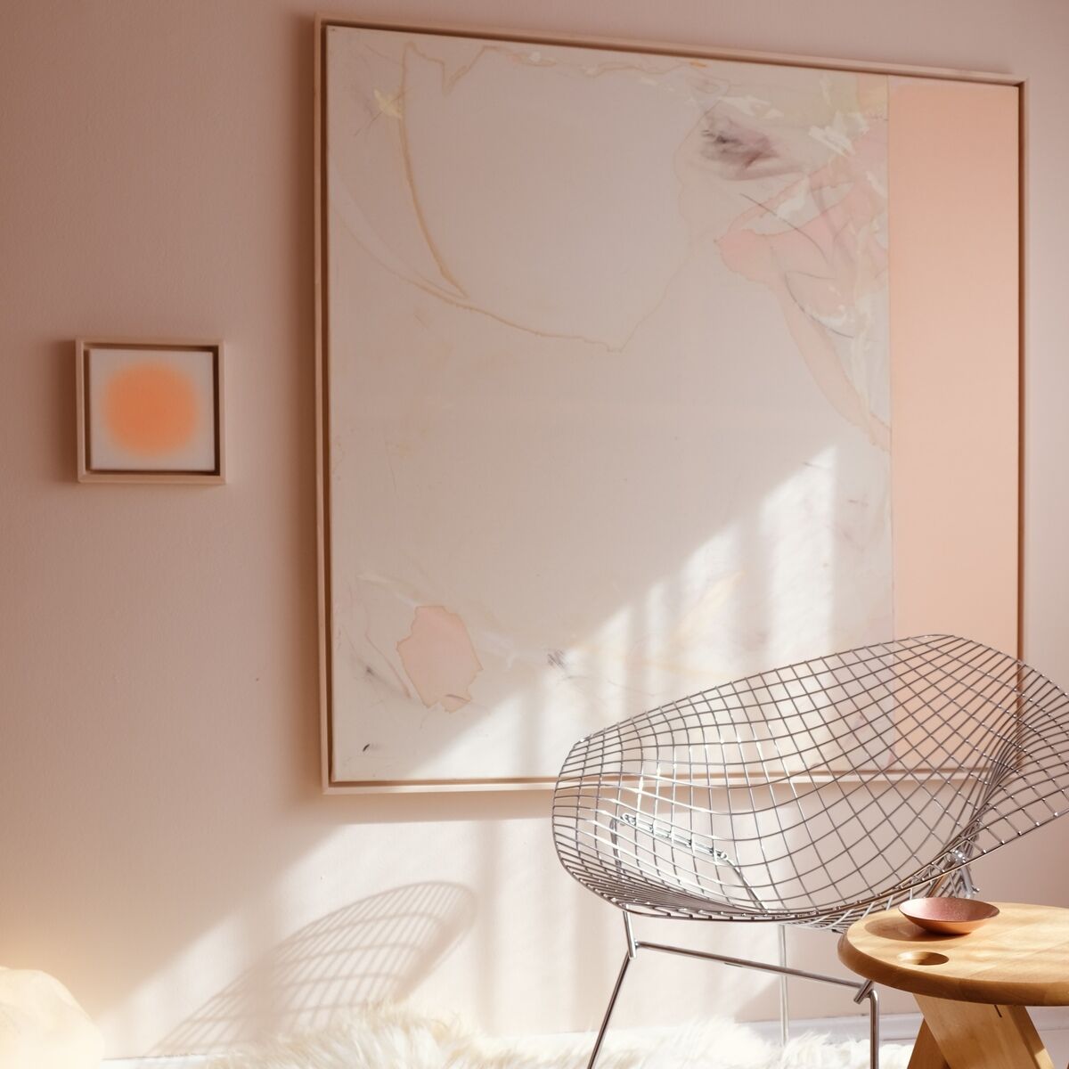 ZEIT-Sonderedition Sorbet »Orange«: Kleines quadratisches abstraktes Kunstwerk mit orangefarbenem Verlauf an einer pastellfarbenen Wand, daneben ein moderner Drahtstuhl und ein heller Fellteppich, daneben ein natürlicher Holztisch; ruhige, minimalistische Wohnraumatmosphäre.