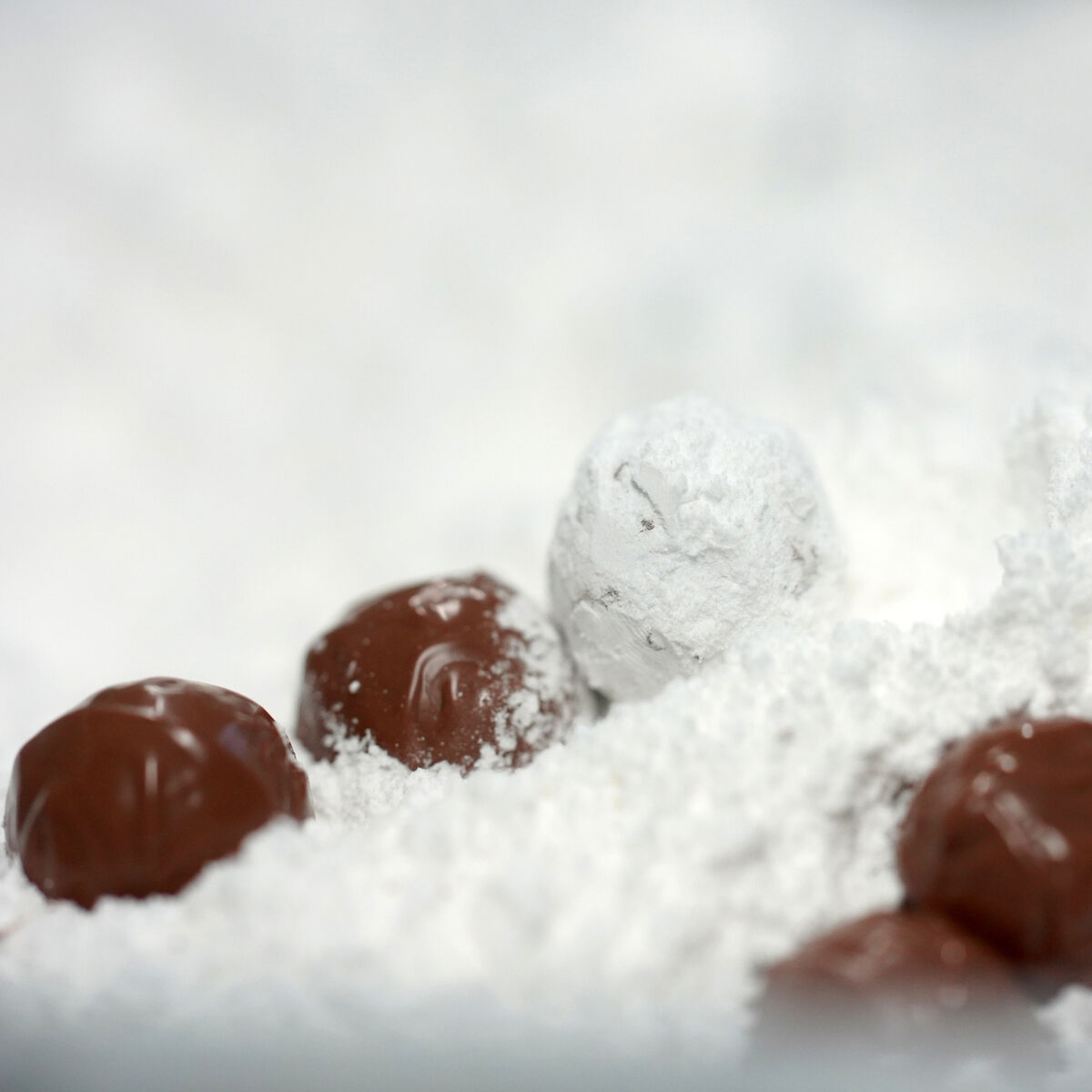 Im Vordergrund liegen mehrere Schokoladentrüffel, teils mit Puderzucker bedeckt. Die cremig-braunen Pralinen liegen auf und in einem weißen Puderzuckerbett. Der Hintergrund ist unscharf, der Fokus liegt auf der feinen Textur und dem Kontrast von brauner Schokolade und weißem Zucker.