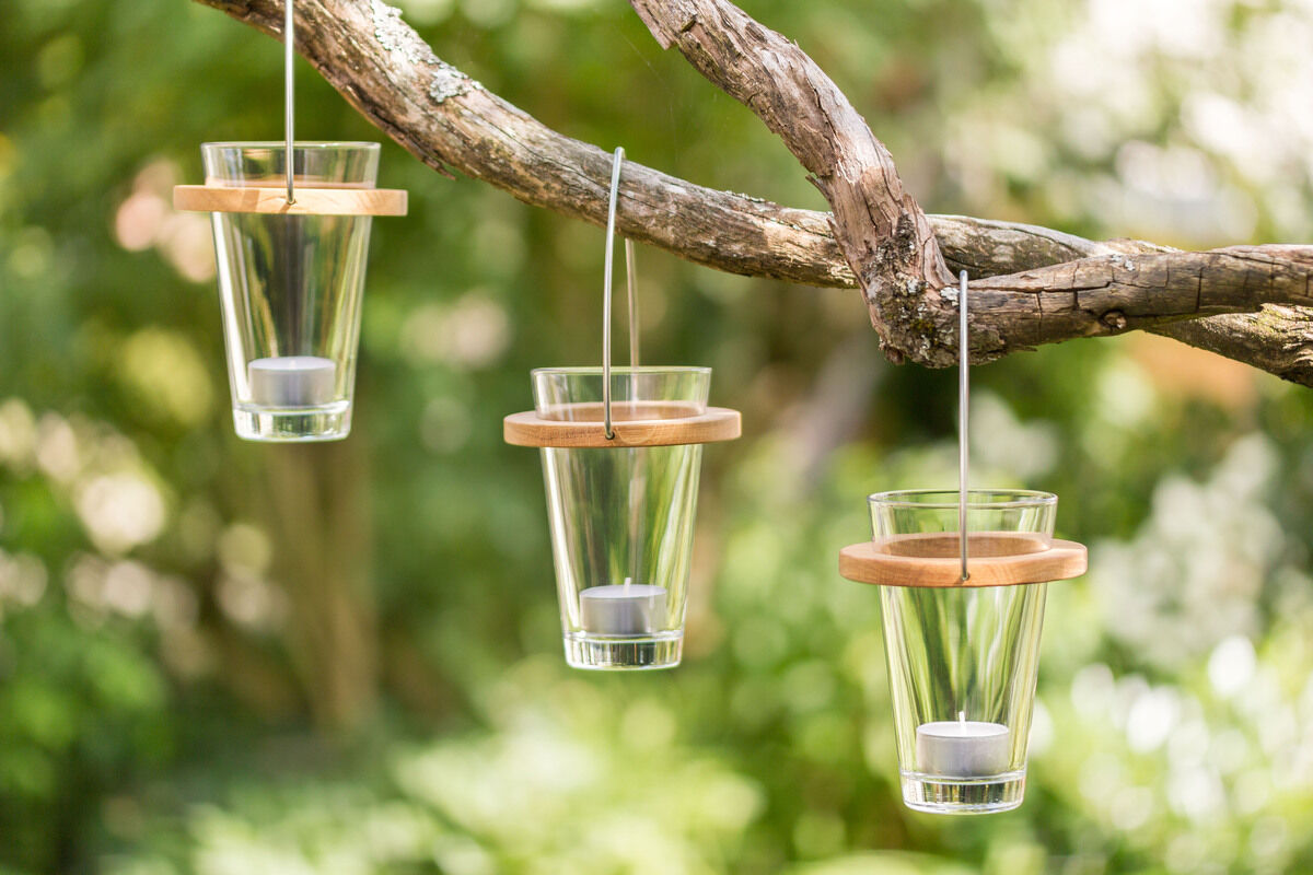 Drei hängende Windlichter aus Glas mit Holzrändern an einem Baumast befestigt, mit weißen Kerzen im Inneren. Hintergrund zeigt unscharfe grüne Pflanzen.