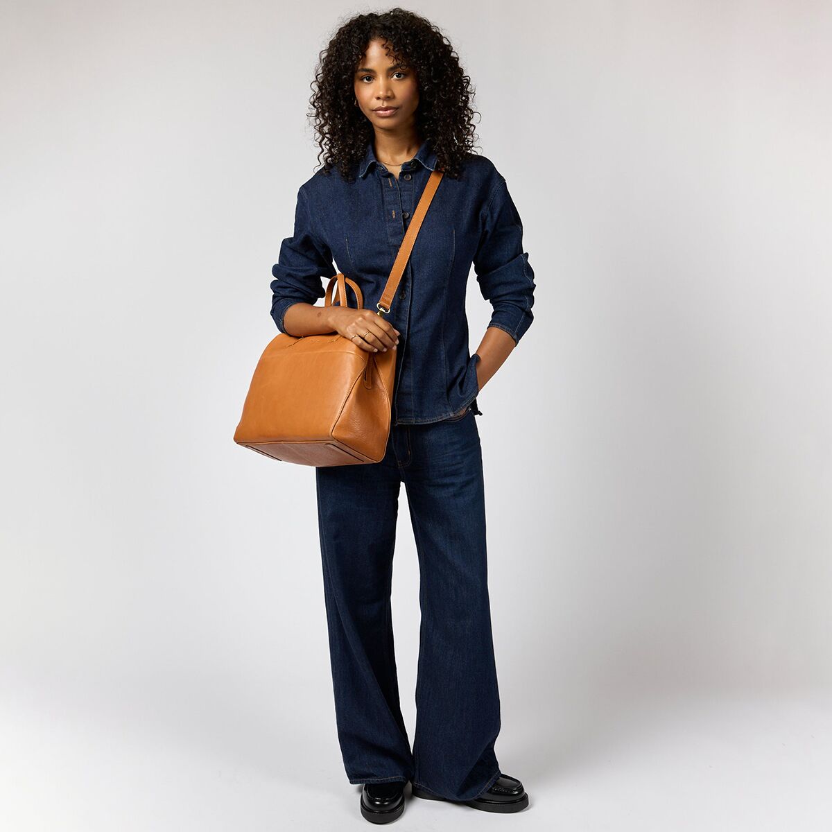 Eine Frau trägt die cognacbraune Schultertasche »Rita« von O My Bag lässig über der Schulter. Sie trägt ein dunkelblaues Jeans-Outfit und schwarze Schuhe. Die geräumige, eckige Ledertasche sticht vor dem neutralen Hintergrund deutlich hervor.