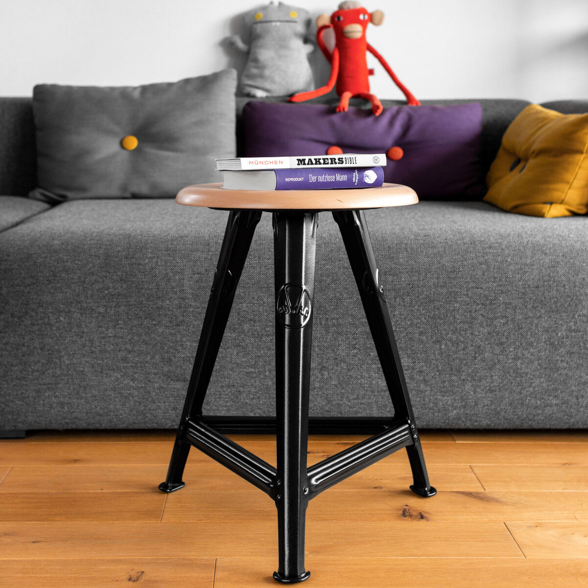 Ein Hocker aus Eichenholz mit mattschwarzen Metallbeinen steht vor einem grauen Sofa. Auf dem Hocker liegen zwei Bücher. Im Hintergrund sind dekorative Kissen und Stofftiere zu sehen.