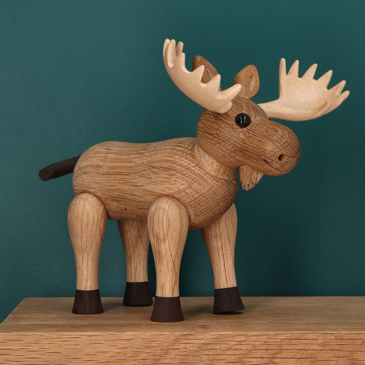 Die Holzfigur »Hirsch Harald« der Marke Spring Copenhagen steht auf einer Holzfläche vor einer petrolfarbenen Wand. Die Figur besteht aus unterschiedlich hellen und dunklen Holztönen, hat detailreiche Geweihpartien, schwarze Beine und Knopfaugen im skandinavischen Design.