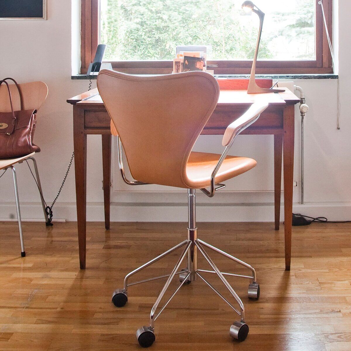 Ein hellbraunes, modernes Home-Office mit dem Schreibtischstuhl »Series 7™« aus langlebigem Essential-Leder von Fritz Hansen im Vordergrund. Der Stuhl hat ein verchromtes Stahlgestell mit fünf Rollen. Am Schreibtisch befinden sich Lampe, Schreibutensilien und ein weiterer gleichartiger Stuhl mit Tasche.
