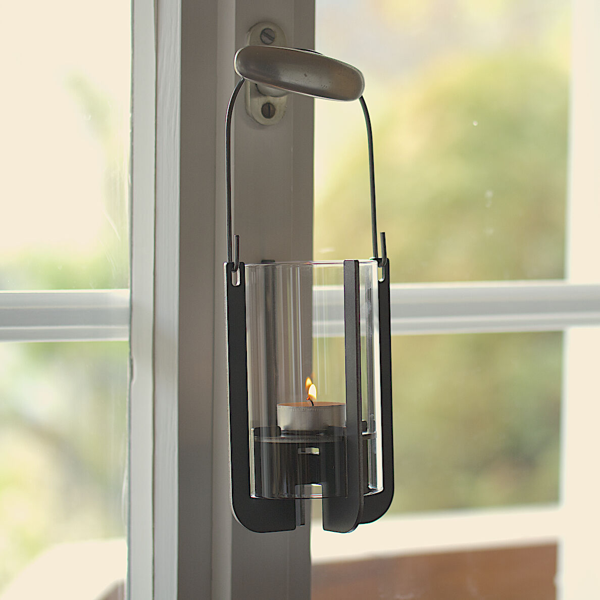 Die Gartenlaterne »Lumi« hängt an einem Türgriff. Sie besteht aus einem modernen, silbernen Halter und einem transparenten Glas, in dem eine brennende Kerze steht. Der Hintergrund zeigt ein Fenster mit Blick auf unscharfe außenliegende Grünelemente.