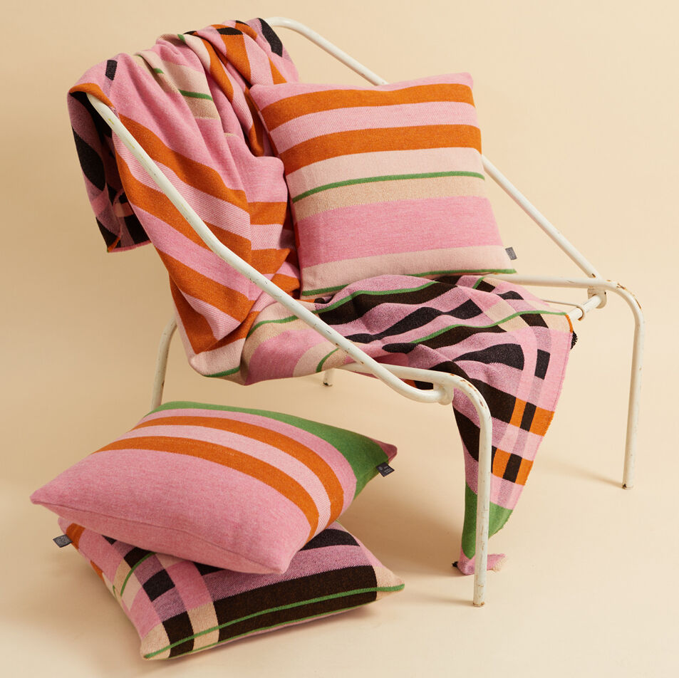 Eine farbenfrohe Decke im Bauhausstil liegt auf einem weißen Stuhl, begleitet von passenden farbenfrohen Kissen auf dem Boden. Die Decke zeigt ein lebendiges Farbblockmuster in Pink, Orange, Grün, Schwarz und Weiß.