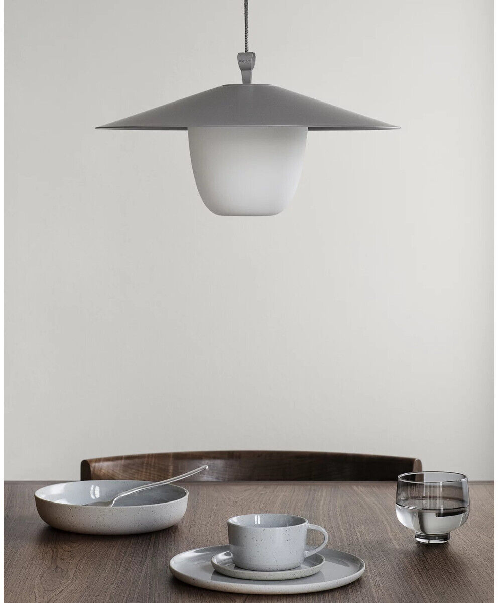 Eine moderne, mobile LED Tischleuchte namens »ANI« in einem minimalistischen Design. Die Lampe steht auf einem Tisch neben einer Tasse, einem Teller und einem Glas. Die Farben sind dezent und bestehen aus Weiß, Grau und Beige.