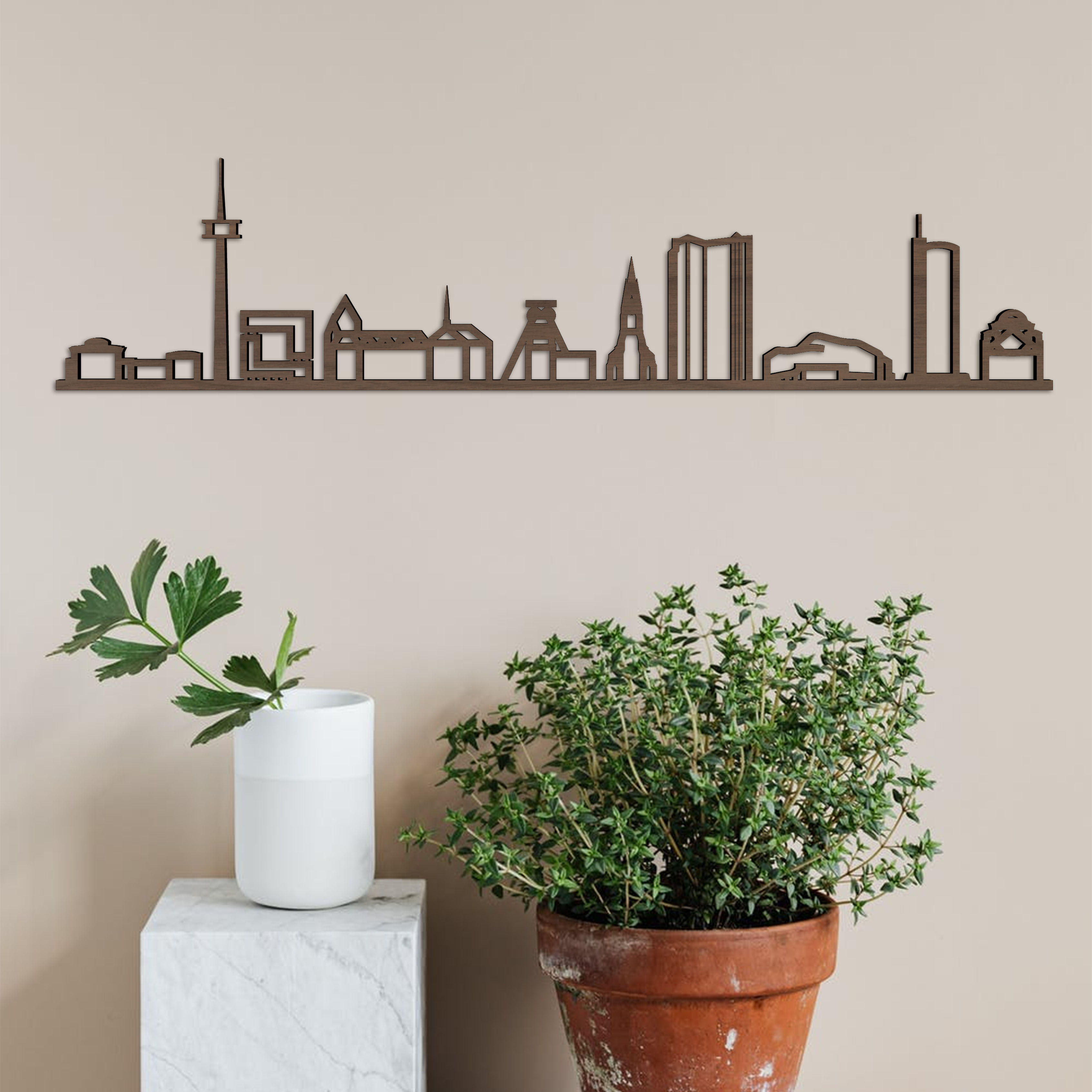 Eine Skyline von Essen aus Holz an einer beige Wand. Darunter stehen eine Pflanze in einem braunen Topf und eine weiße Vase mit grünen Blättern auf einem weißen Podest.