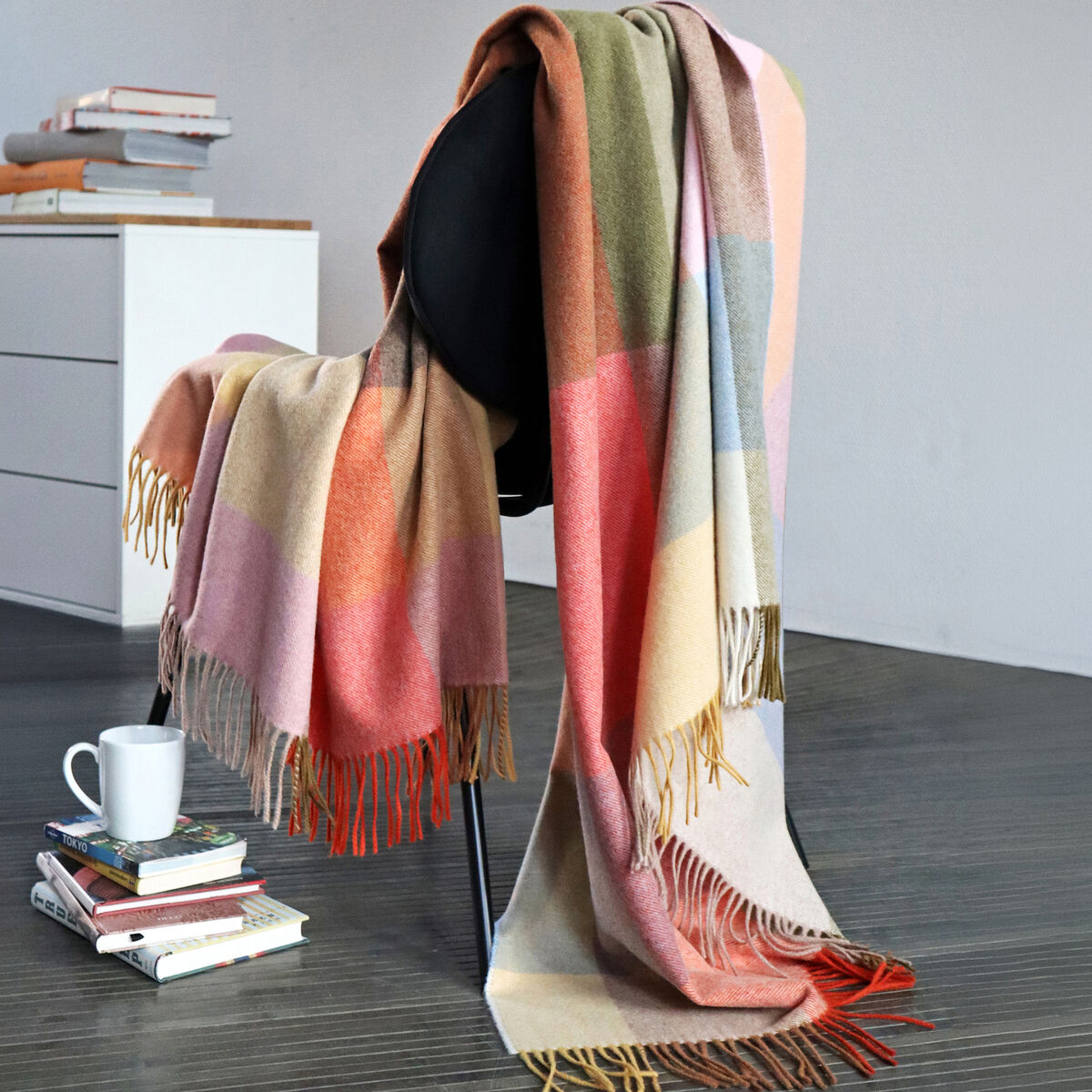Merinowolldecke »Rimini« von Eagle Products in modernen, hellen Farben mit asymmetrischem Karo-Design liegt dekorativ über einem schwarzen Stuhl. Die weiche Wolldecke hat Fransen, daneben Bücherstapel und eine weiße Tasse auf dem Holzboden im stilvollen Wohnraum.