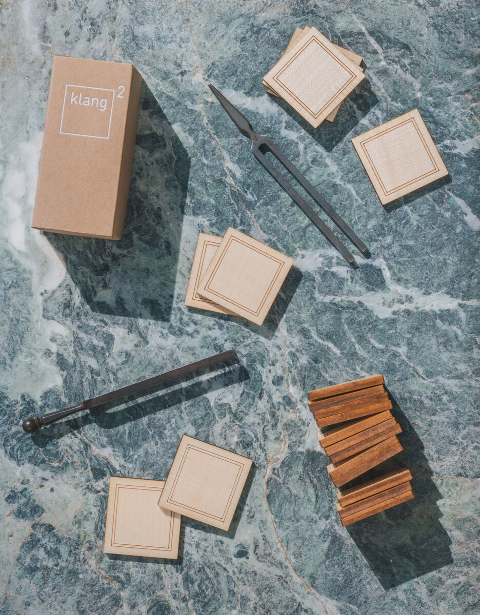 Spielesammlung »Geigenbauer« von klang² bestehend aus mehreren braunen Holzplättchen, zwei Werkzeugen und einer Kartonverpackung auf hell- und dunkelgrauer Marmoroberfläche.