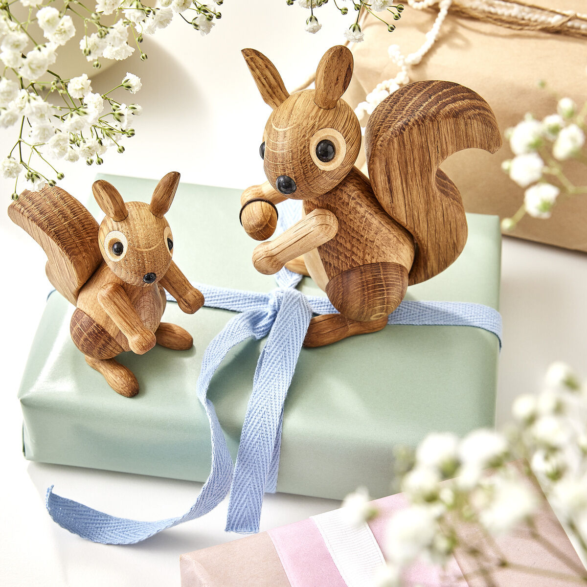 Zwei unterschiedlich große Eichhörnchen-Holzfiguren sitzen auf liebevoll verpackten Geschenken, verziert mit blauen und weißen Schleifenband. Im Vorder- und Hintergrund sind weiße Blüten zu sehen, die eine frühlingshafte und festliche Atmosphäre schaffen.