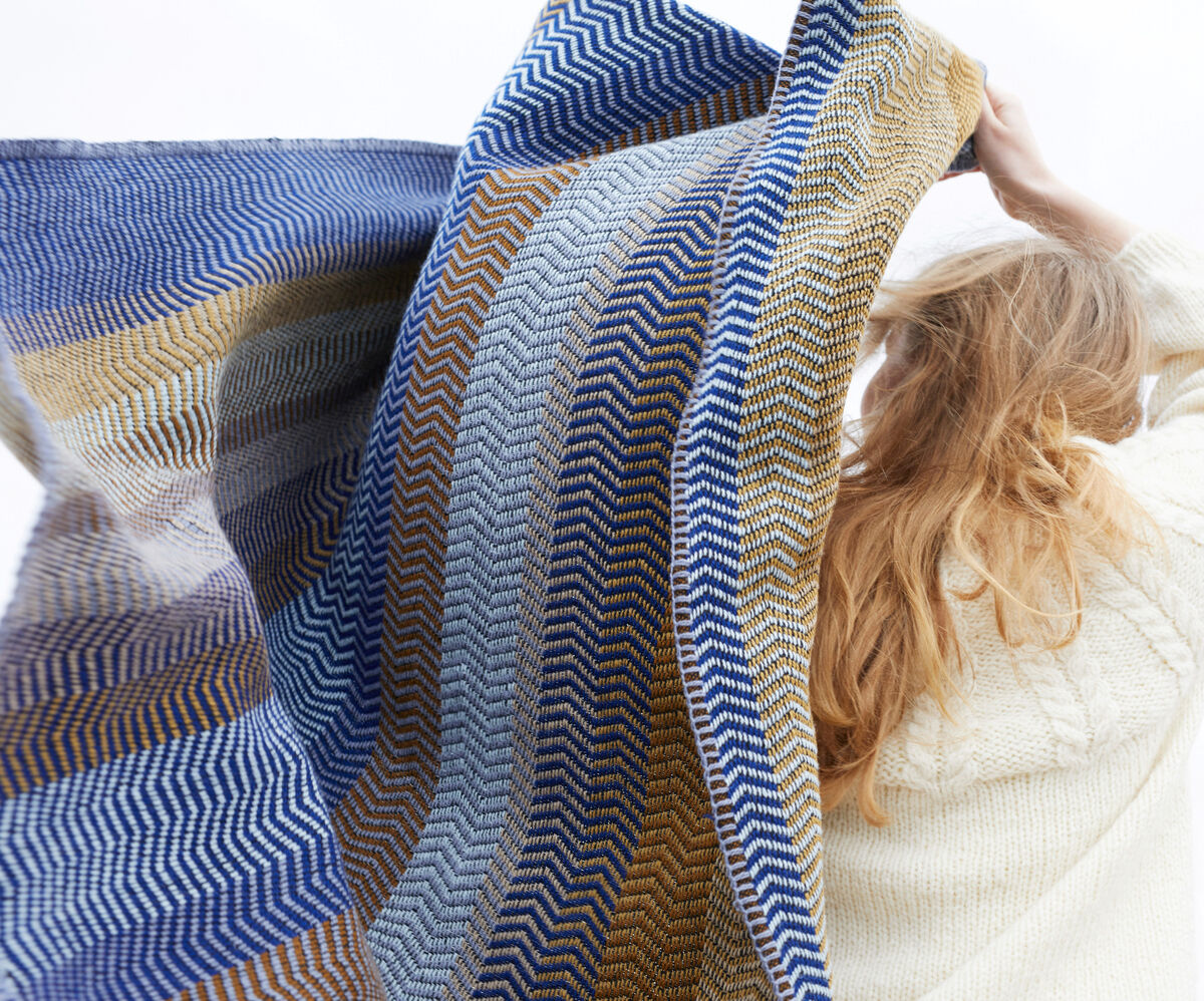 Eine Frau in einem beigen Pullover hält die Lammwolldecke »Fri« hoch. Die Decke hat ein Zickzackmuster in Blau-, Braun- und Gelbtönen.