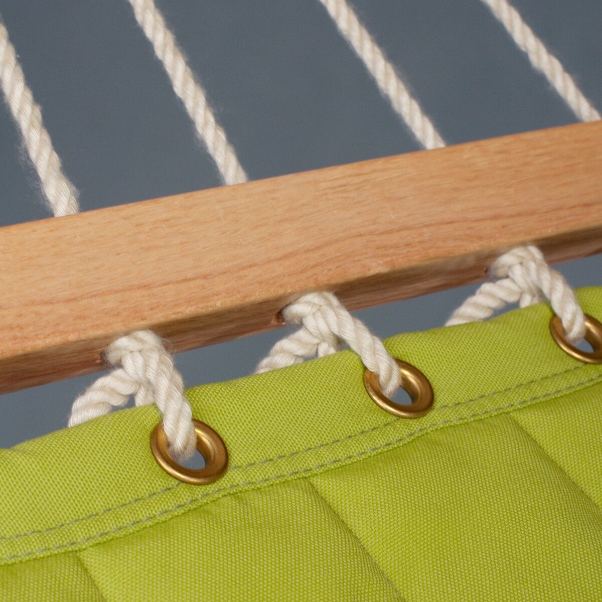 Nahaufnahme einer grünen Hängematte mit hölzernem Spreizstab, weißen Seilen und robusten goldenen Ringen. Die Hängematte hängt vor einem blau-grauen Hintergrund.