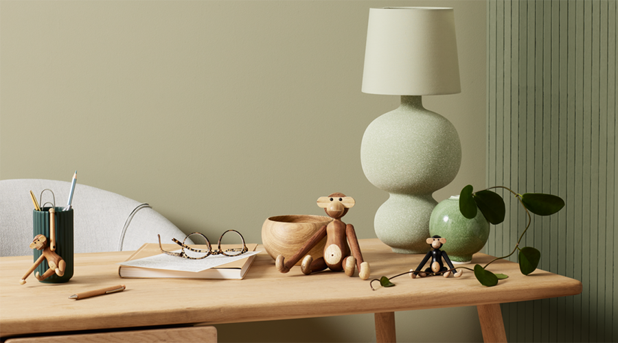Holztisch mit zwei Holzfiguren von Affen, Holzschale, Brille auf Buch, Stifthalter mit Stiften, grüne Tischlampe und grüne Pflanze vor beiger und grüner Wand