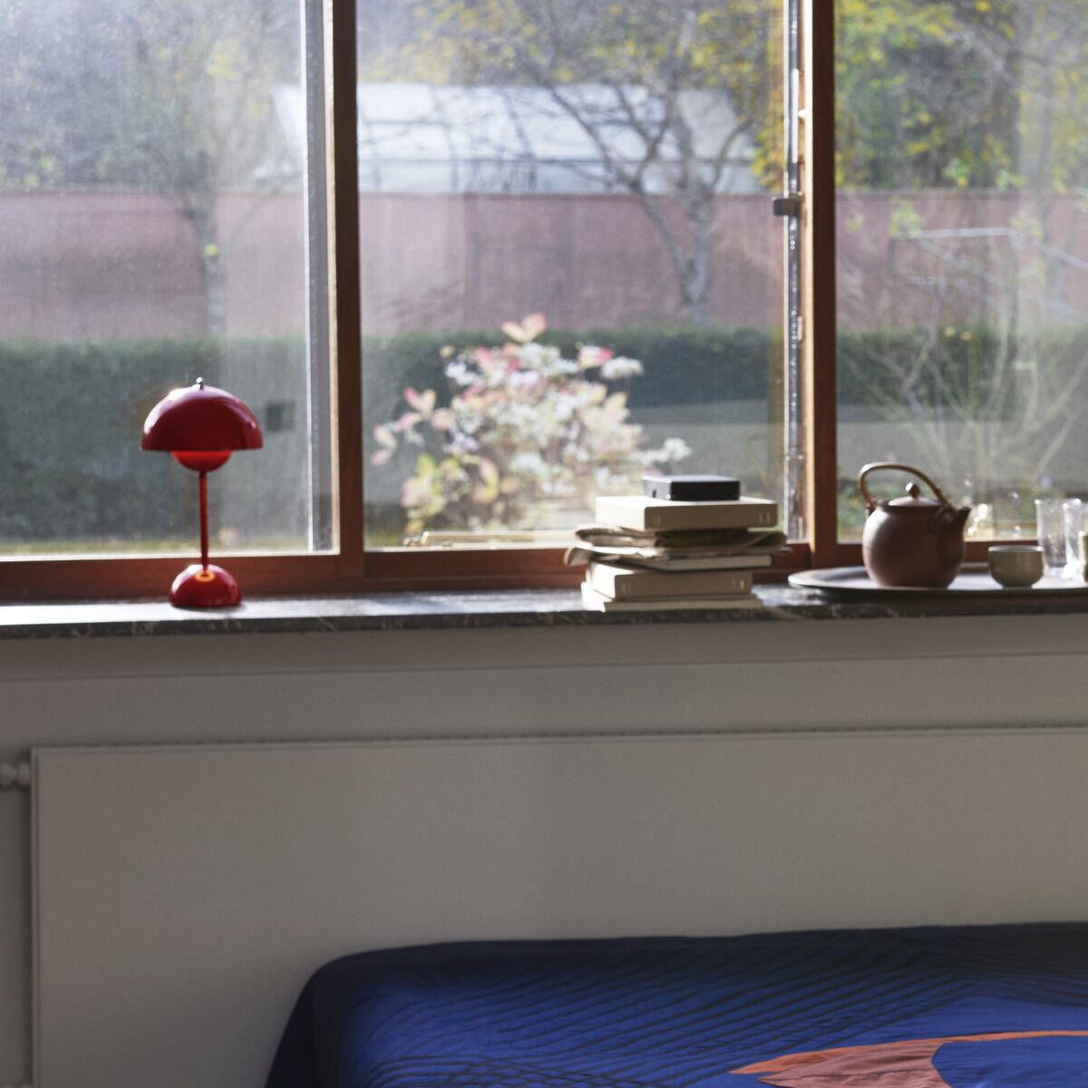 Eine Flowerpot VP9 Tischleuchte Akku in Rot steht auf einer Fensterbank neben einem Stapel Bücher und einer Teekanne. Das Zimmer hat ein großes Fenster mit Blick auf einen Garten. Ein Bett mit einer blauen Decke befindet sich im Vordergrund.