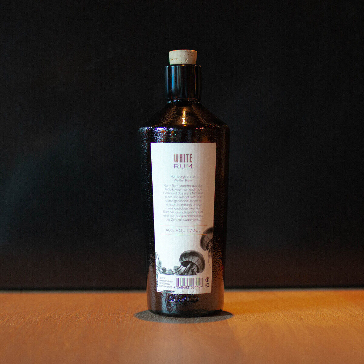 Das Bild zeigt eine Flasche mit der Aufschrift 'WHITE RUM' auf einem Etikett. Die Flasche steht vor einem dunklen Hintergrund auf einer Holzoberfläche. Die Flasche hat einen metallischen Glanz und ist mit einem Korken verschlossen.