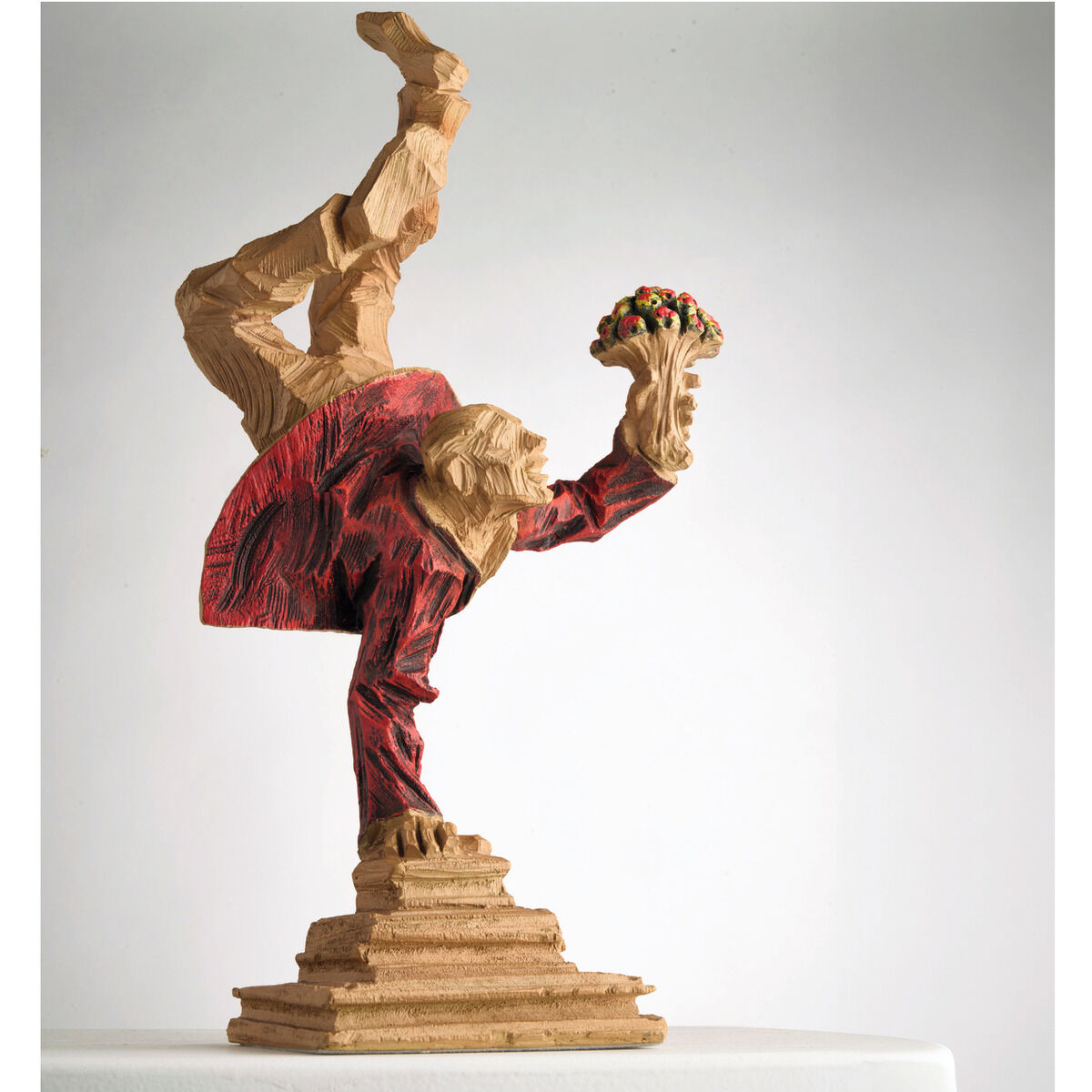 Eine Skulptur in Holzoptik von Roman Johann Strobl, dargestellt in einer dynamischen Pose mit einem Blumenstrauß in der Hand. Die Figur trägt ein rotes Outfit und balanciert auf einem sockelförmigen Podest.