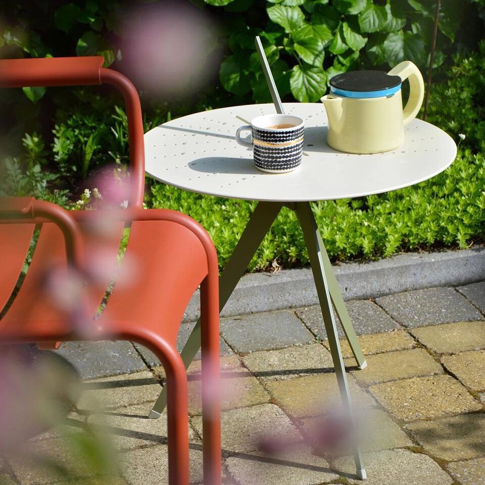 Ein moderner, kleiner Tisch mit dem Namen »Sundial« auf einer Terrasse, umgeben von grünen Pflanzen. Auf dem Tisch stehen eine Tasse und eine Kanne. Neben dem Tisch steht ein roter Stuhl. Der Boden besteht aus Pflastersteinen in Naturtönen.